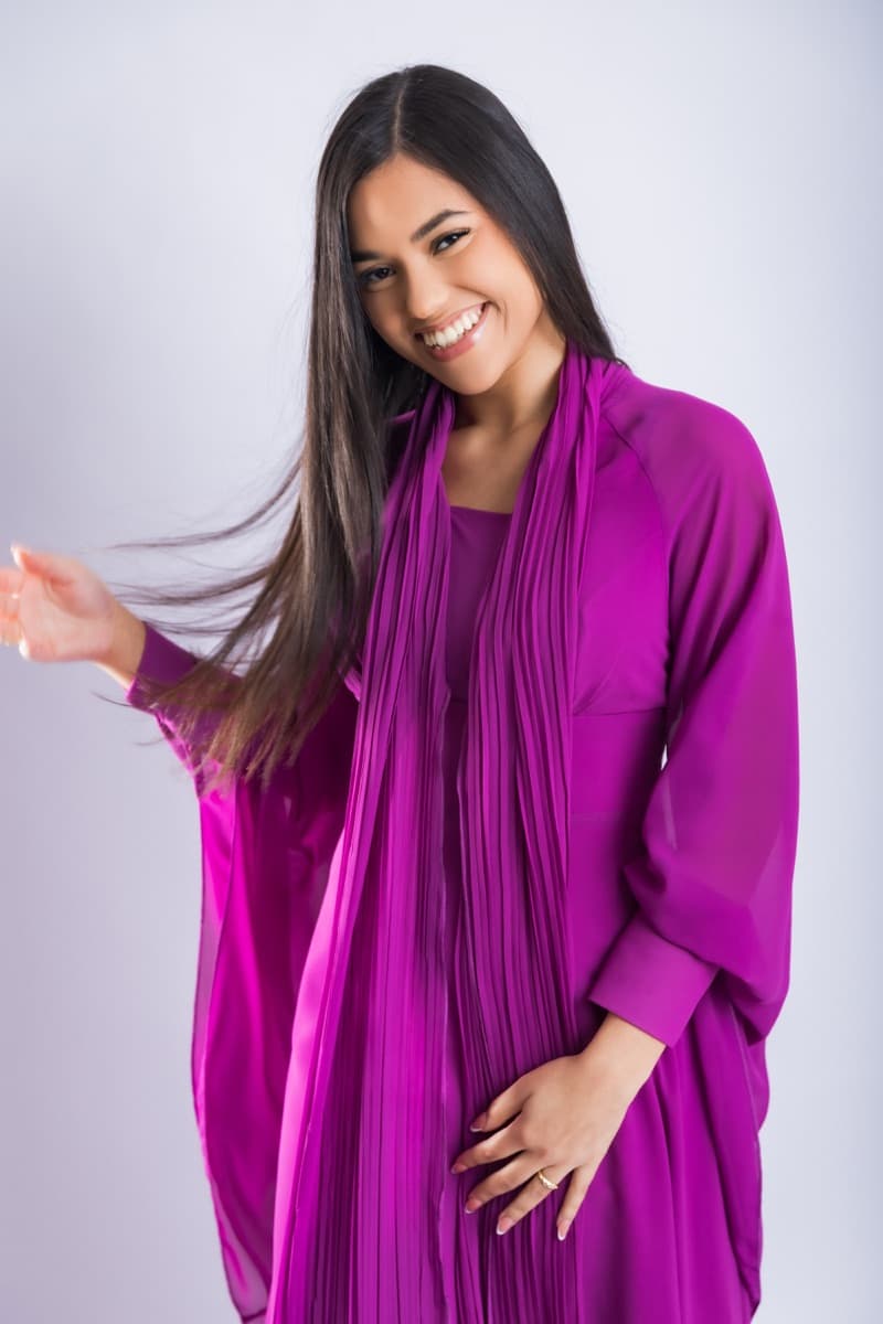 Elizabet Maria sonriendo, con vestido fucsia plisado y el brazo extendido